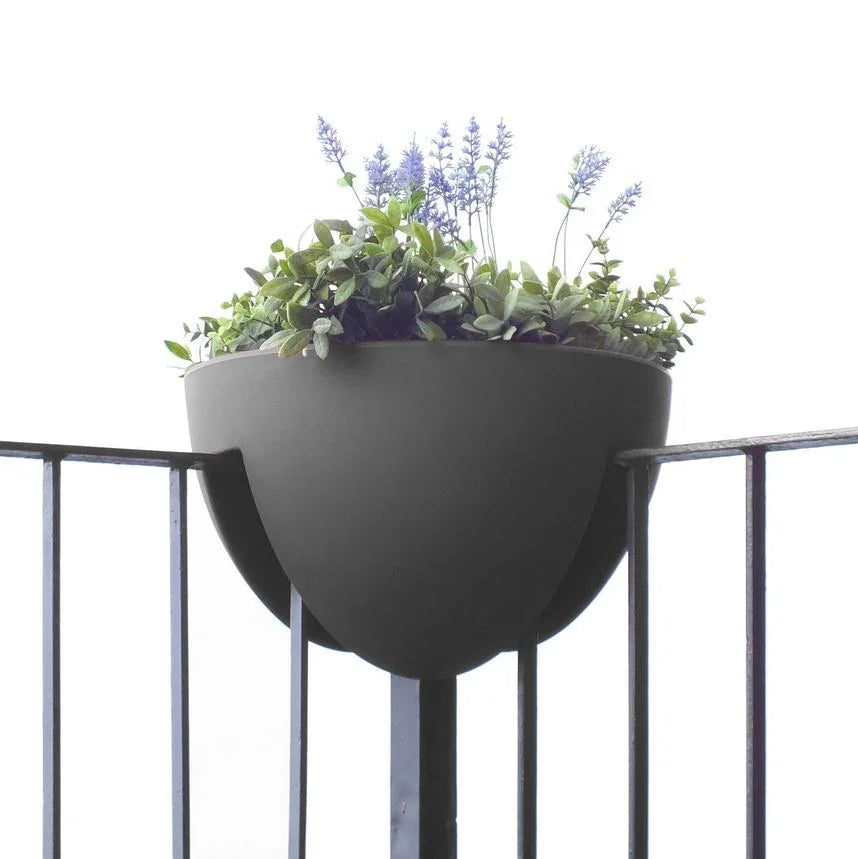 Ein moderner, schwarzer Wandpflanzer mit üppigen grünen Pflanzen und violetten Blumen, montiert auf einem Balkongeländer vor einem weißen Hintergrund.