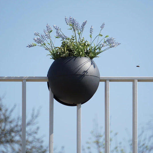 Ein runder, schwarzer Blumentopf mit violetten Blumen hängt über einem weißen Metallgeländer vor einem klaren, blauen Himmel; in der Nähe ist ein kleines Insekt zu sehen.