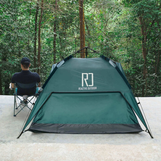 Eine Person sitzt in einem Campingstuhl neben einem grünen Zelt mit der Aufschrift „REACTIVE OUTDOOR“, umgeben von üppigen Bäumen in einer ruhigen Waldlandschaft.