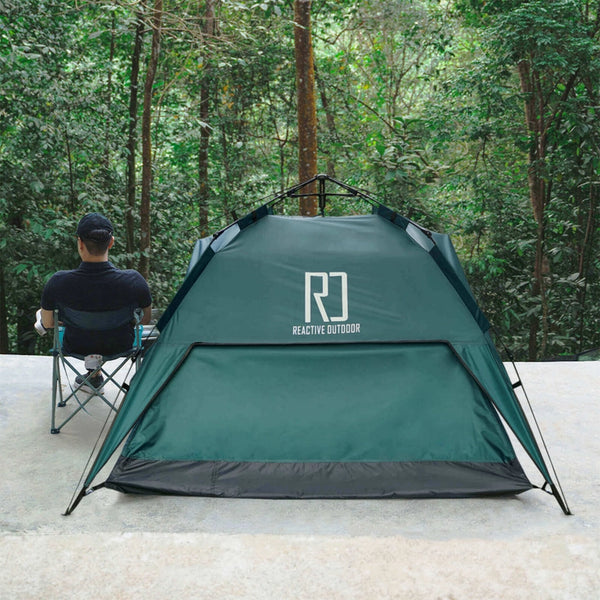 Eine Person sitzt in einem Campingstuhl neben einem grünen Zelt mit der Aufschrift „REACTIVE OUTDOOR“, umgeben von üppigen Bäumen in einer ruhigen Waldlandschaft.