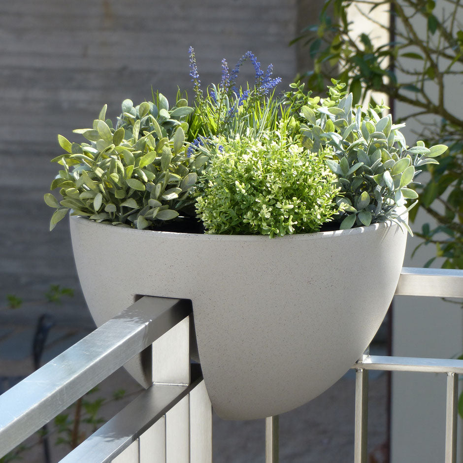 Ein moderner, runder Blumentopf, gefüllt mit üppigen grünen Kräutern und kleinen violetten Blumen, aufgehängt an einem Balkongeländer.